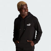 Puma Essential Small Logo Overhead Fleece Hoodie in black, front view worn by a model showing a ribbed hood, a small Puma Cat Logo on the chest, and a kangaroo pocket.