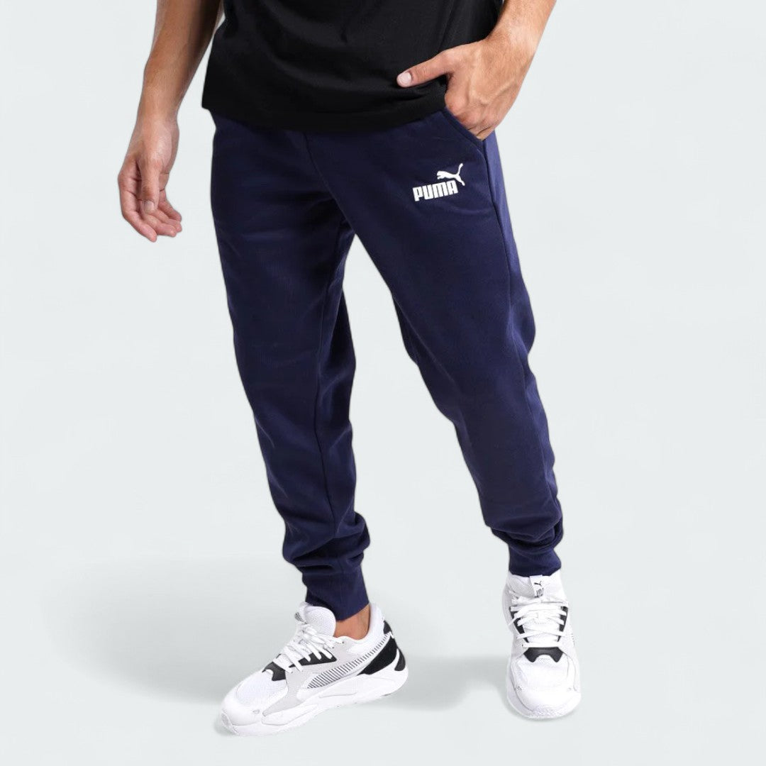 Puma Essential Logo Pants Men's in navy fleece, front view worn by a model showing the elastic waistband, drawcord, and a white Puma Cat Logo on the thigh.