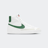 Nike Blazer Mid '77 Juniors' trainers in white and green accents, side profile showing classic mid-cut leather upper, and large green Nike Swoosh.