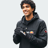 A hand wearing the Ellesse Daxo Touchscreen Glove in black, demonstrating its snug fit and the ability to use a smartphone with the glove on.