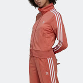 Side profile of the Adidas Firebird track top in Magic Earth brown, showing the classic white 3-Stripes running from the shoulder to the ribbed cuff.