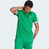 Model profile of the green Adicolor Sport t-shirt, showcasing the iconic white 3-Stripes running from the shoulder to the sleeve hem.