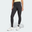 Front three-quarter view of a model wearing the black adidas Neuclassics leggings paired with white trainers, showing the high-waisted fit, full-length leg coverage, and three horizontal white stripes wrapping around the left lower shin.
