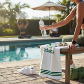 Person by a pool holding an Adidas towel and water bottle, with another person exercising in the background.