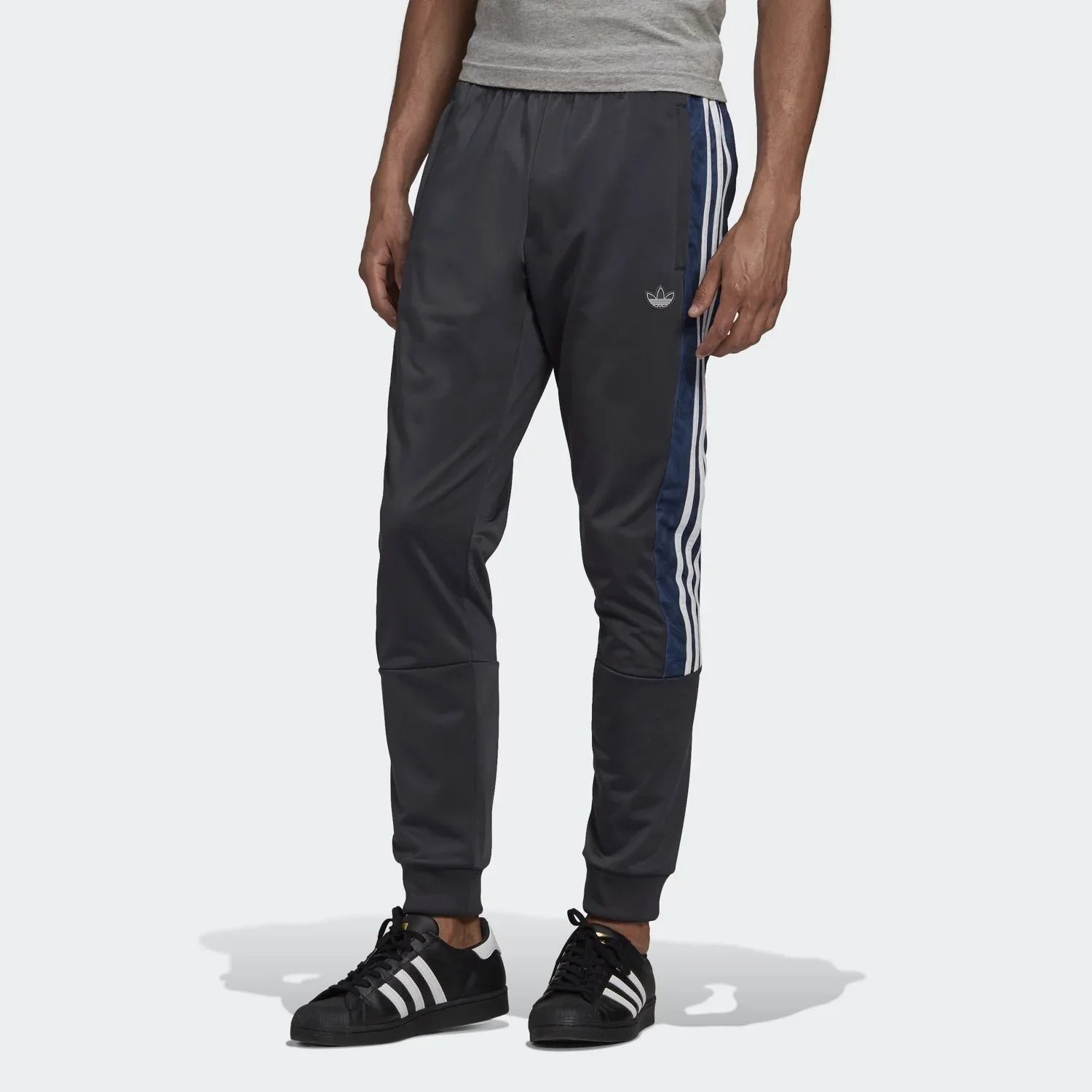 A man wearing Adidas men's slim-fit graphic sweat pants in night grey color with blue stripes on the side, paired with a grey crew neck t-shirt and black and white sneakers.