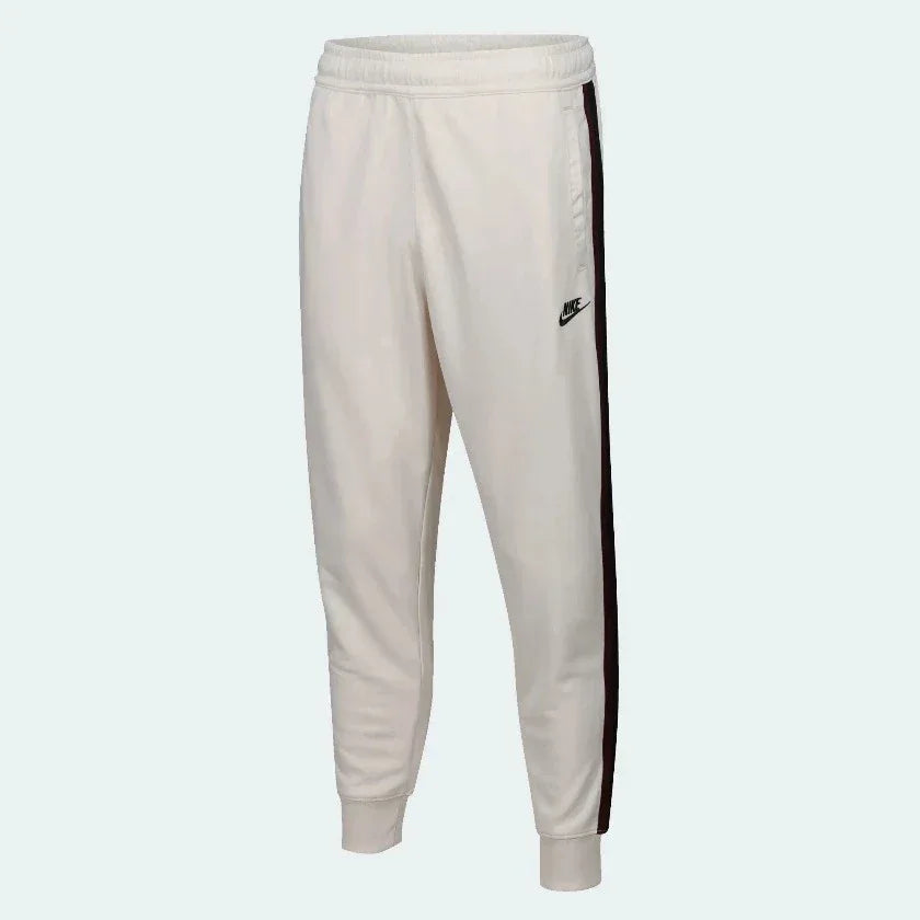 Nike Men's Tribute Woven Track Pants in light beige, front view showing elastic waistband, white side stripes/piping, and Nike Swoosh logo.