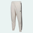 Nike Men's Tribute Woven Track Pants in light beige, front view showing elastic waistband, white side stripes/piping, and Nike Swoosh logo.