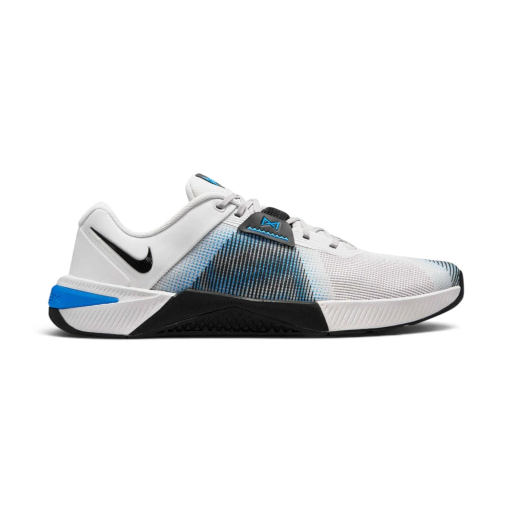 Medial side profile of the Nike Free Metcon 10 men's training shoe in a light grey and white mesh upper, showcasing the extended black rubber rope-wrap on the inner midsole, a blue lace-lock tab accent, a small black Swoosh on the collar, and a vibrant blue heel clip. Main image