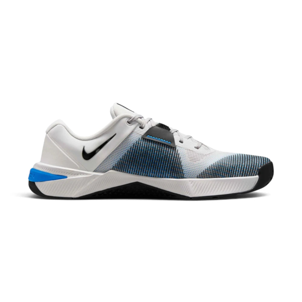 Lateral side profile showing the full silhouette of the shoe, featuring a light grey breathable upper, black side wrap overlay, black Nike Swoosh logo on the rear white panel, and a sculpted white midsole resting on a black rubber outsole with a bright blue heel clip.