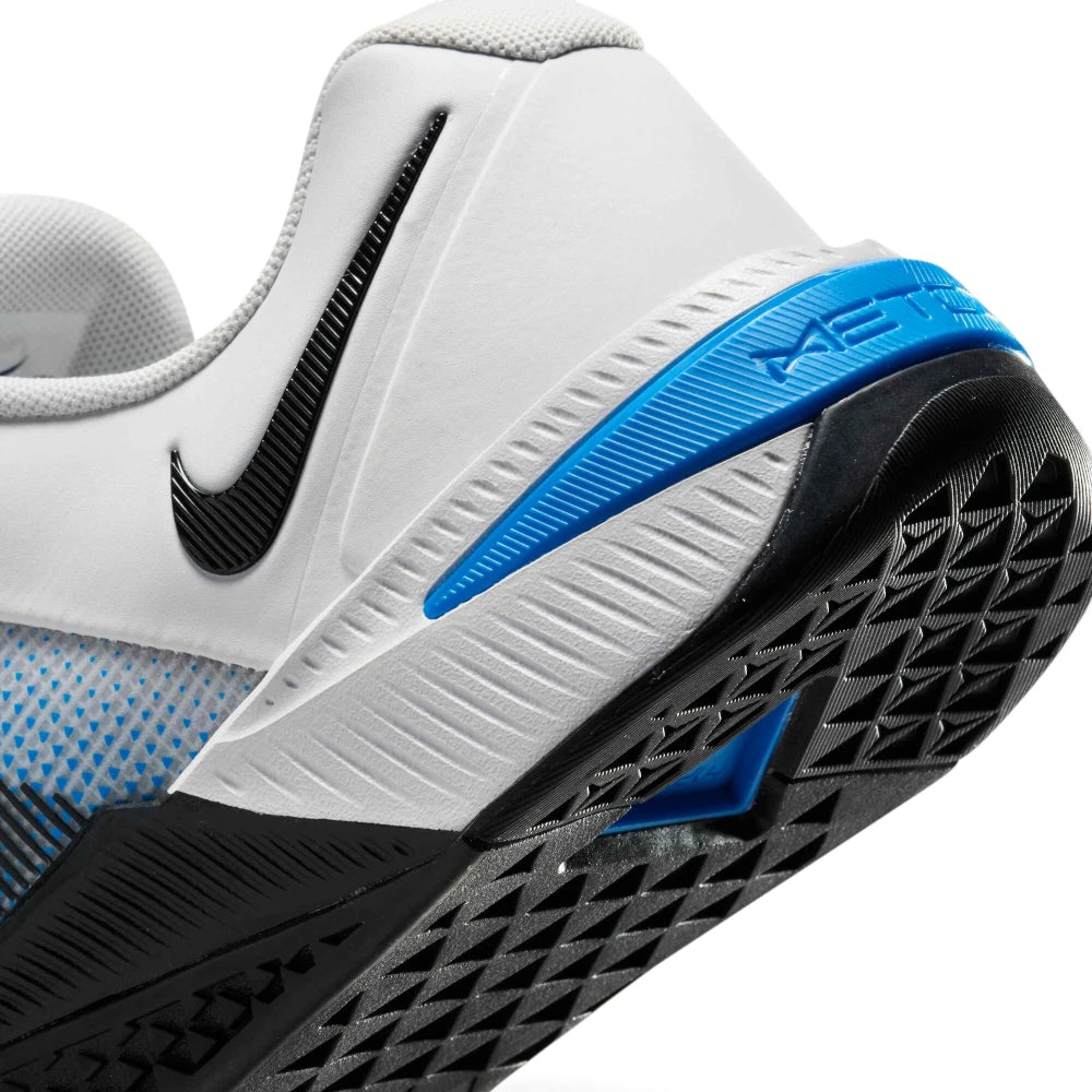 Close-up shot focusing on the rear outsole and heel construction, showing the grooved white foam midsole, black rubber outsole with diamond-pattern tread, black Swoosh, and the blue Metcon heel clip.