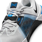 Close-up view of the forefoot and lacing system, displaying the light grey mesh texture, integrated laces, black textured midfoot overlay, and the black lace-lock tab with a blue Metcon logo and blue "Lace Tuck" tongue tab.