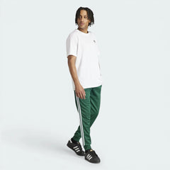 The Collegiate Green SST pants, highlighting the slim fit and the high-contrast white 3-Stripes running down the leg.