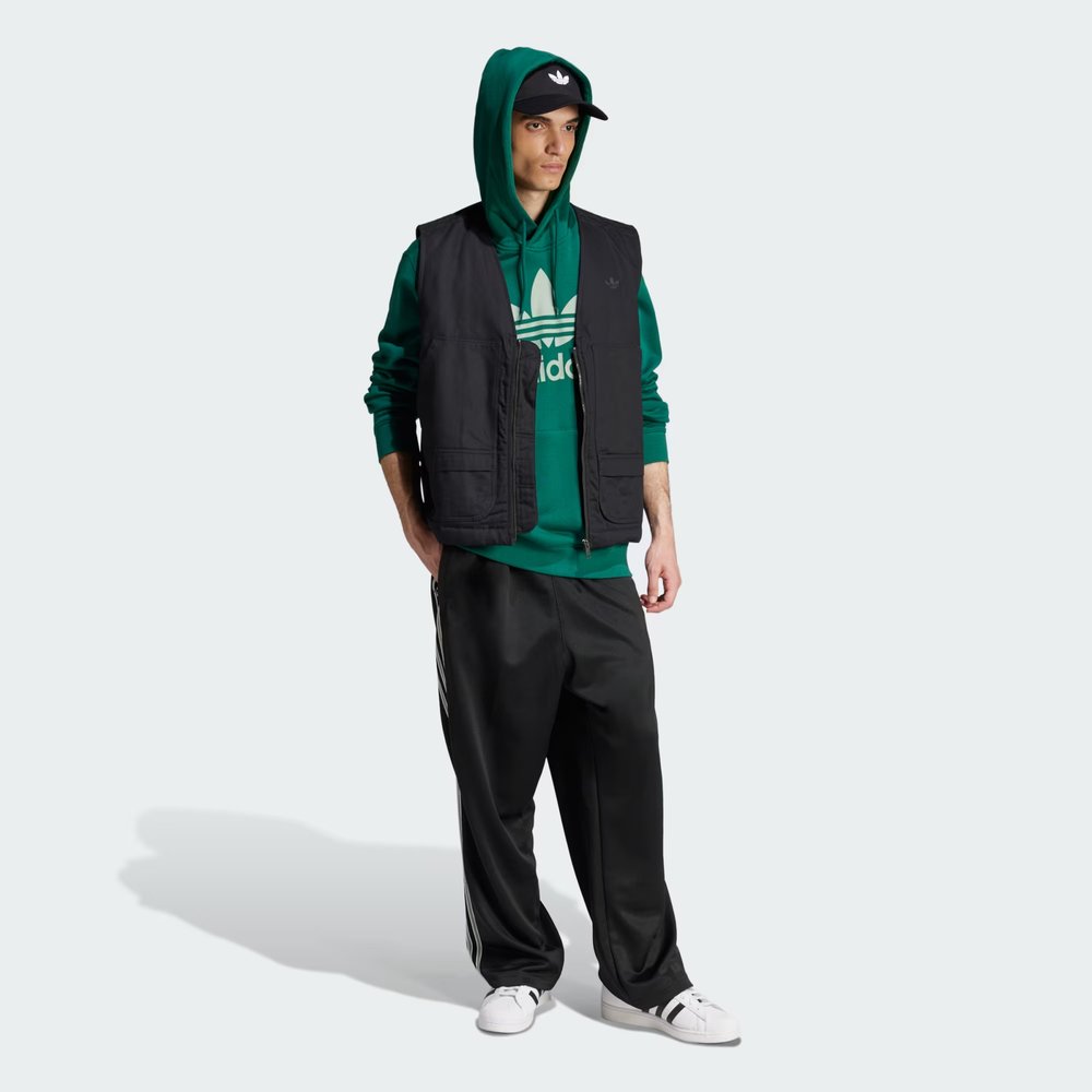 Person wearing a green hoodie, black vest, and black pants with white sneakers on a white background