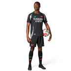 Full-length studio shot of a male model wearing the complete away kit, including the black authentic jersey, matching shorts with red accents, and black socks featuring team branding.