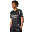 Studio lifestyle shot of a model wearing the Arsenal 24/25 away jersey in black, highlighting the flat-knit V-neck collar in Pan-African flag colours, white Emirates Fly Better sponsor logo, green adidas logo, and intricate side panel graphics.