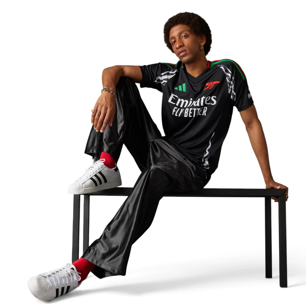 Full-length studio shot of a model seated on a bench wearing the black jersey paired with satin track trousers and white Superstar trainers, showcasing its everyday streetwear versatility.