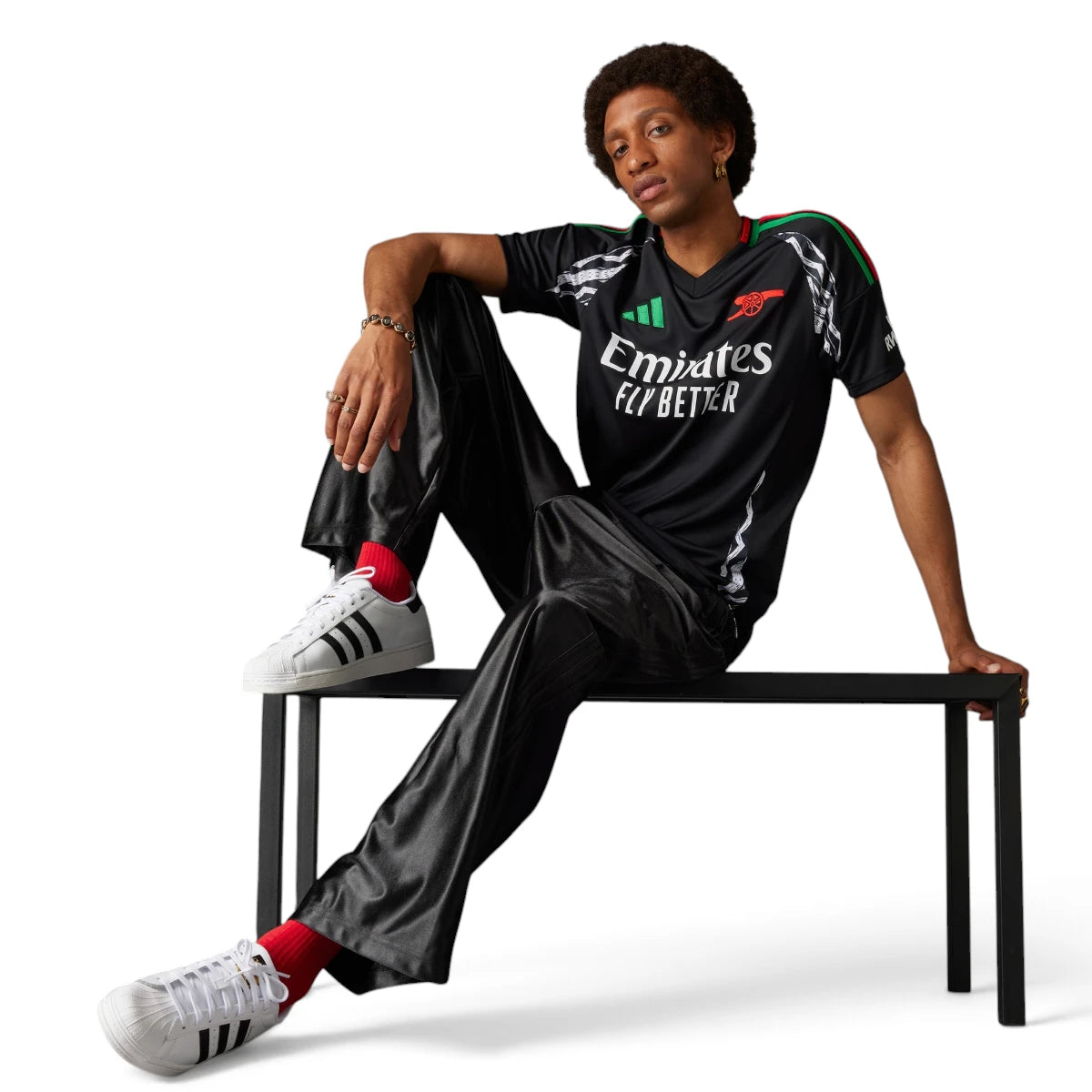 Full-length studio shot of a model seated on a bench wearing the black jersey paired with satin track trousers and white Superstar trainers, showcasing its everyday streetwear versatility.