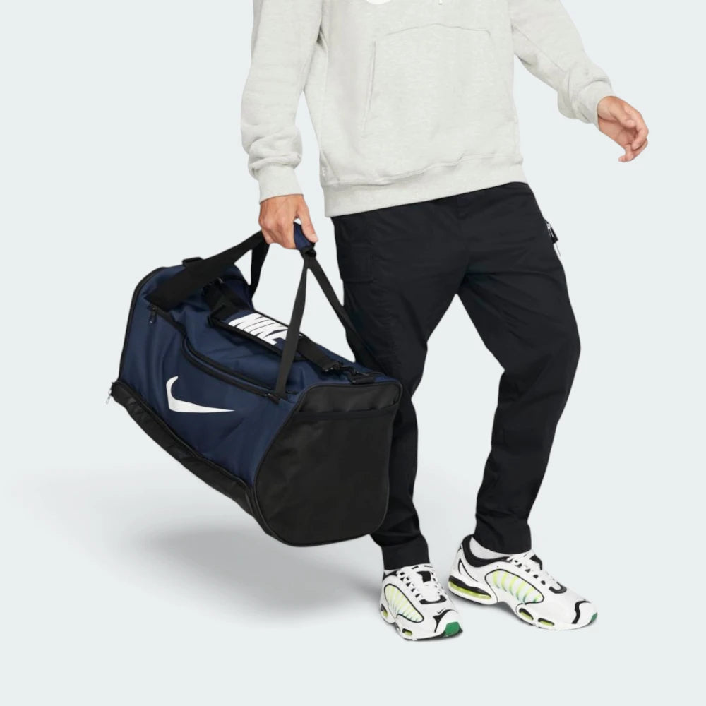 Demonstrates the duffel bag being carried by hand using the padded dual grab handles, showing the proportion and scale of the 60L medium duffel against an athlete in athletic wear.