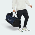 Demonstrates the duffel bag being carried by hand using the padded dual grab handles, showing the proportion and scale of the 60L medium duffel against an athlete in athletic wear.