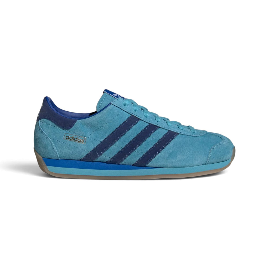 Side Profile Image: Studio lateral shot of the adidas Country Japan in Preloved Blue, highlighting the pigskin nubuck upper, darker blue serrated three stripes, gold foil adidas stamp, and semi-translucent gum outsole wrapping up the heel.   