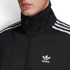 Close-up zoom on the fabric of the Adidas Firebird Track Jacket in black, showing the smooth tricot texture and the embroidered white Trefoil logo detail on the chest.