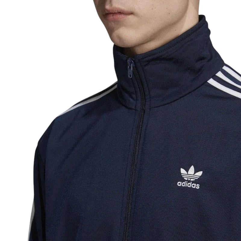 Close-up zoom on the Adidas Firebird Track Jacket Men's, showing the smooth tricot fabric texture and the embroidered white Trefoil logo detail on the chest.