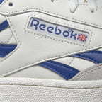 Close-up macro view of the lateral leather upper, showcasing the embossed Reebok window box logo with the Union Jack flag, royal blue leather stripe accents, fine contrast stitching, and textured midsole rim.