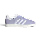 Displays the full lateral silhouette featuring the soft Violet Tone suede upper, contrasting Cloud White serrated 3-Stripes, gleaming metallic gold foil "Gazelle" lettering, matching white heel tab, and the clean rubber cupsole.