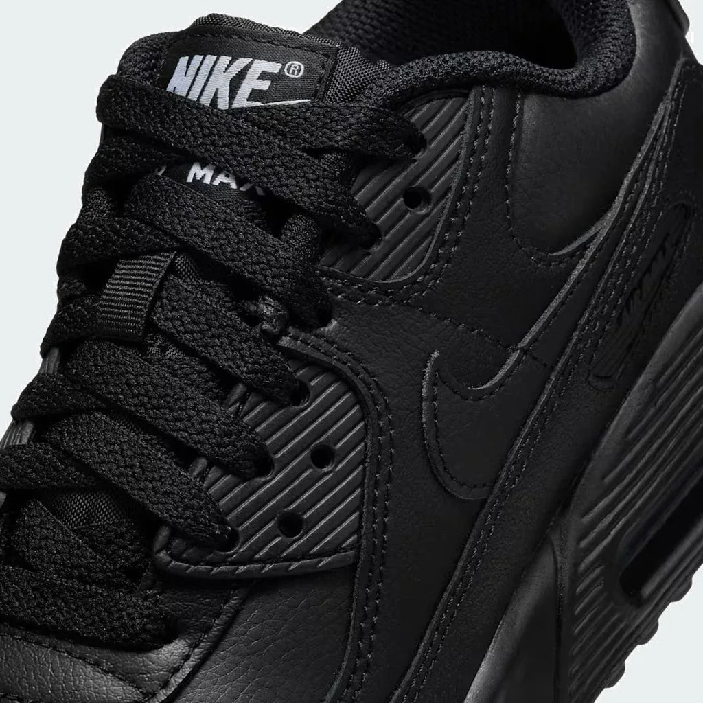 Close-up zoom on the front of the Nike Air Max 90 Juniors' black trainers, showing the mesh toe box, lacing system, and layered synthetic overlays.