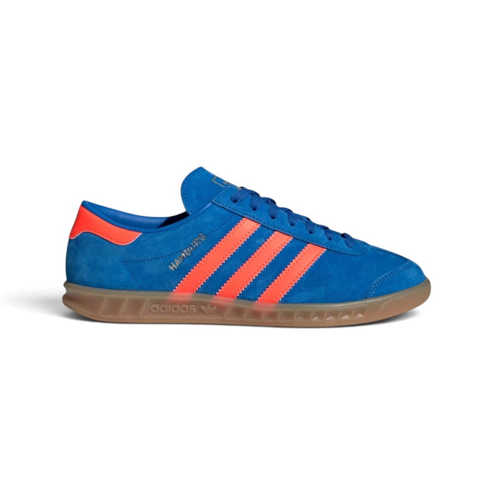 Highlights the sleek low-profile lateral silhouette featuring the rich Bluebird suede upper, vibrant Semi Solar Orange leather serrated 3-Stripes, metallic gold foil "HAMBURG" script, matching orange heel tab, and semi-translucent Gum 3 rubber cupsole with debossed adidas Trefoil branding. Main image