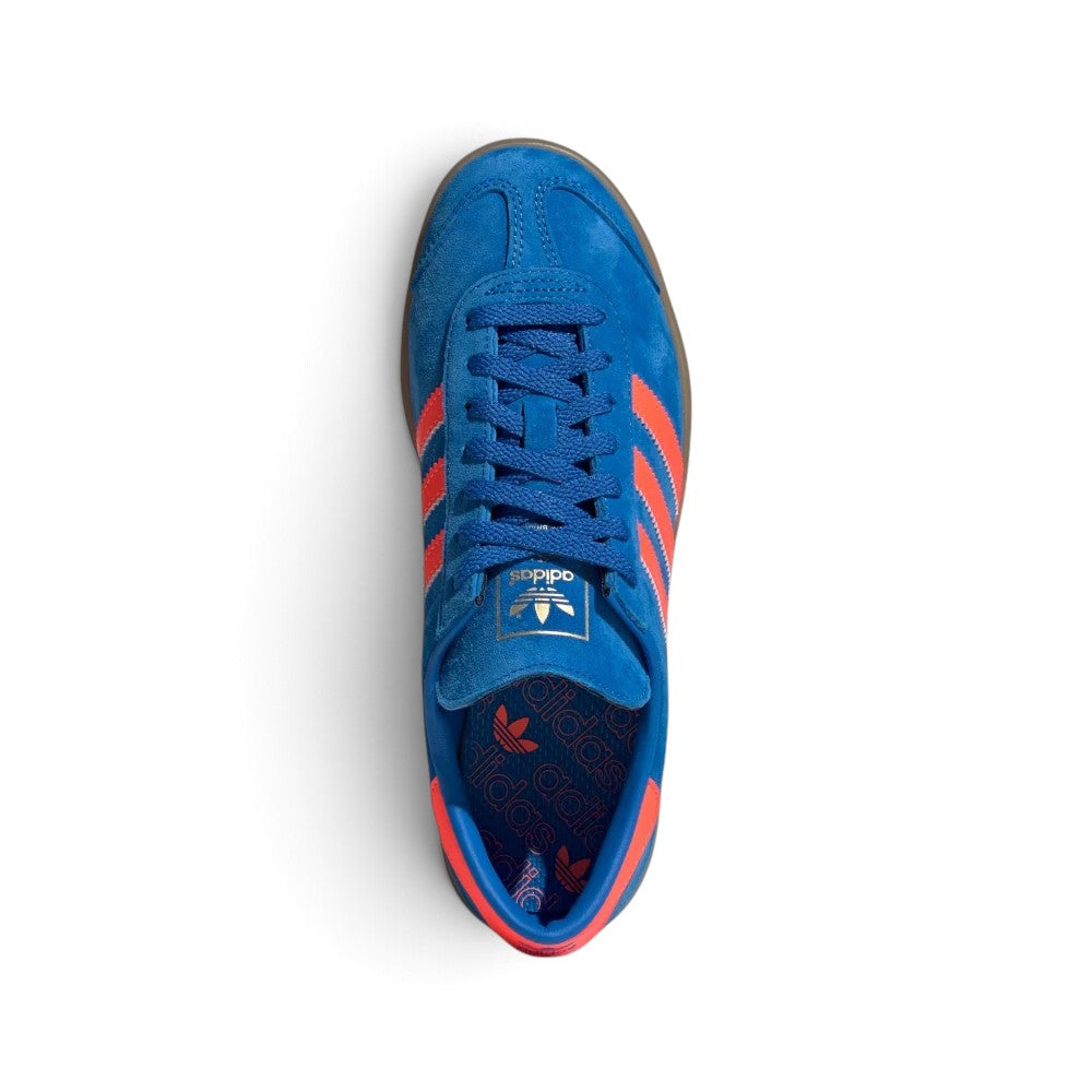 Focuses on the locked-in central lacing layout, displaying flat Bluebird blue laces, thin suede tongues stamped with gold foil adidas Trefoil logo badges, reinforced T-toe overlays, and royal blue insoles patterned with repeating orange Trefoil graphics.