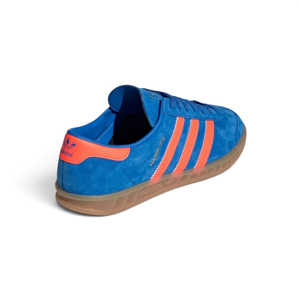 Focuses on the rear three-quarter profile, displaying the vibrant Semi Solar Orange leather heel moustache tab stamped with a blue Trefoil logo, clean heel counter stitch lines, and the textured rim of the translucent gum cupsole.