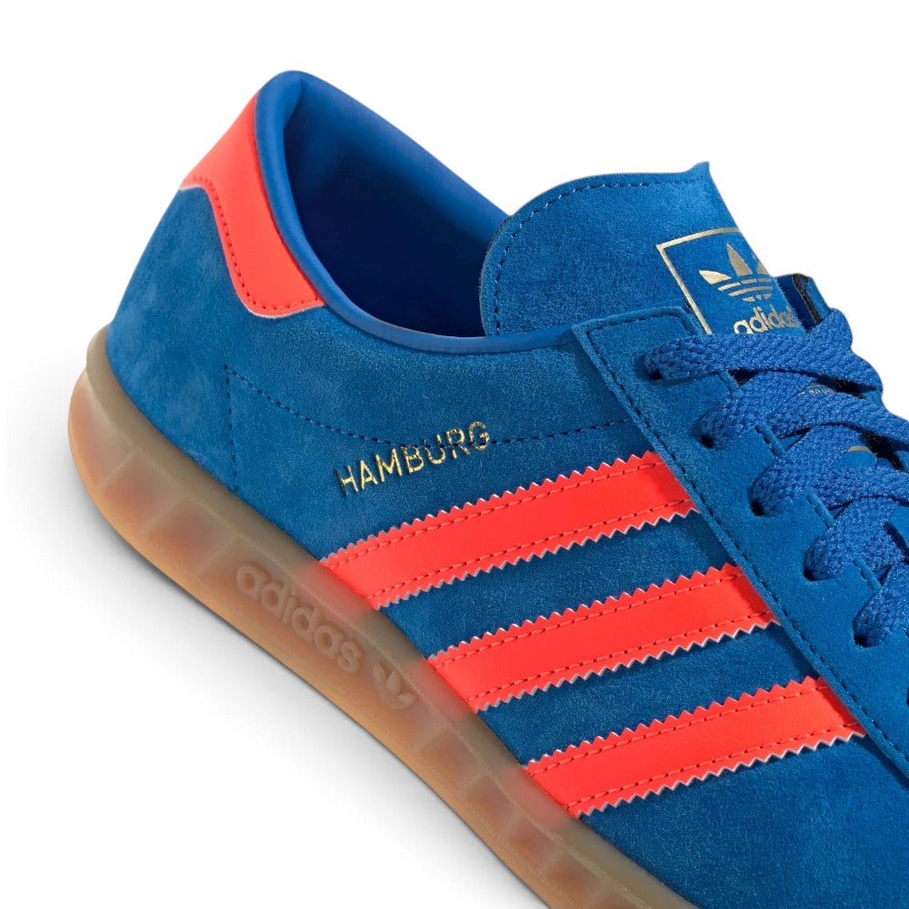 Details the micro-precision of the midfoot and lacing area, showing the gilded metallic "HAMBURG" wordmark, crisp zig-zag edges of the orange serrated 3-Stripes, tonal eyelets, and the gold foil Trefoil tongue crest.