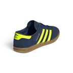 Focuses on the rear three-quarter profile, displaying the vibrant Solar Yellow leather heel moustache tab stamped with an indigo Trefoil logo, clean heel counter stitch lines, and the textured rim of the translucent gum cupsole.