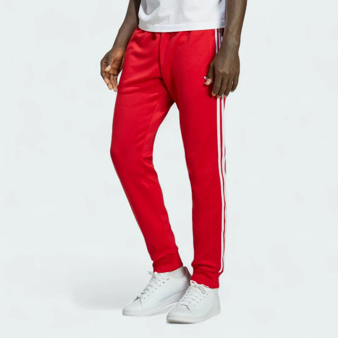 Adidas Superstar Track Pants Men's in red, front view showing white 3-Stripes down the legs, Trefoil logo on the hip, and zipped pockets. Main image