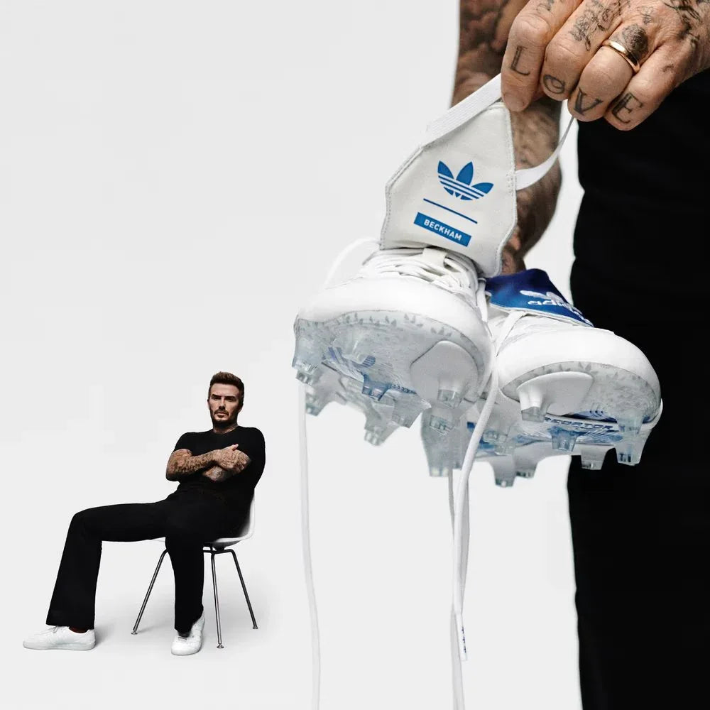 Person holding a pair of Adidas soccer cleats with David Beckham sitting on a chair in the background.