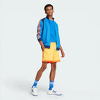 Person wearing a blue jacket with orange stripes, yellow shorts, and white sneakers on a light gray background