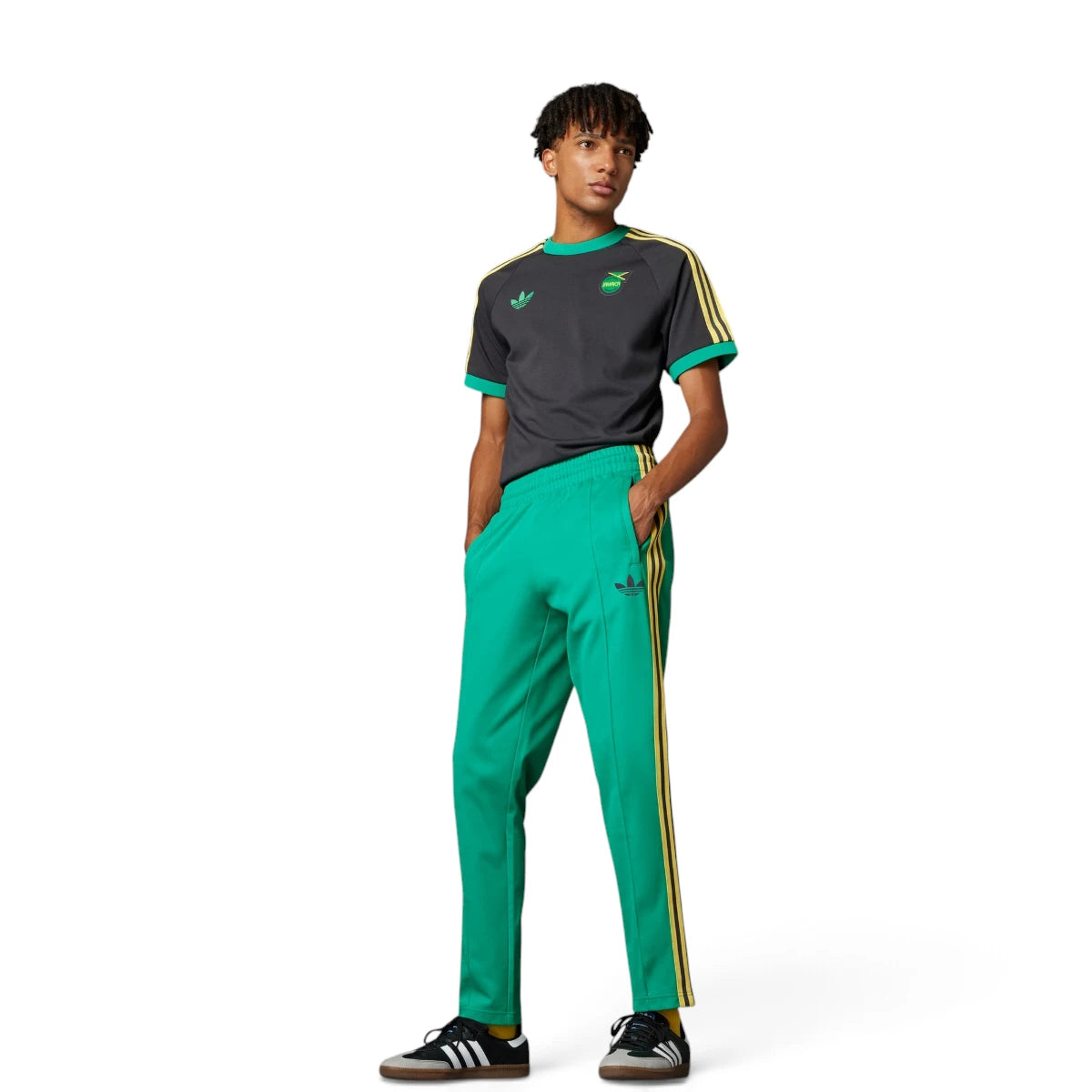 Full-body studio shot of a model wearing the adidas Jamaica Beckenbauer Track Pants in Court Green, paired with a dark grey t-shirt and black trainers, highlighting the slim fit, yellow 3-Stripes taping, and the Jamaica football federation crest.