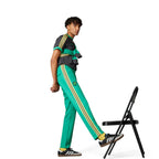 Studio action view of a model in a dynamic seated stretch pose lifting one leg toward a chair, demonstrating the flexibility and slim-fit comfort of the green track trousers.