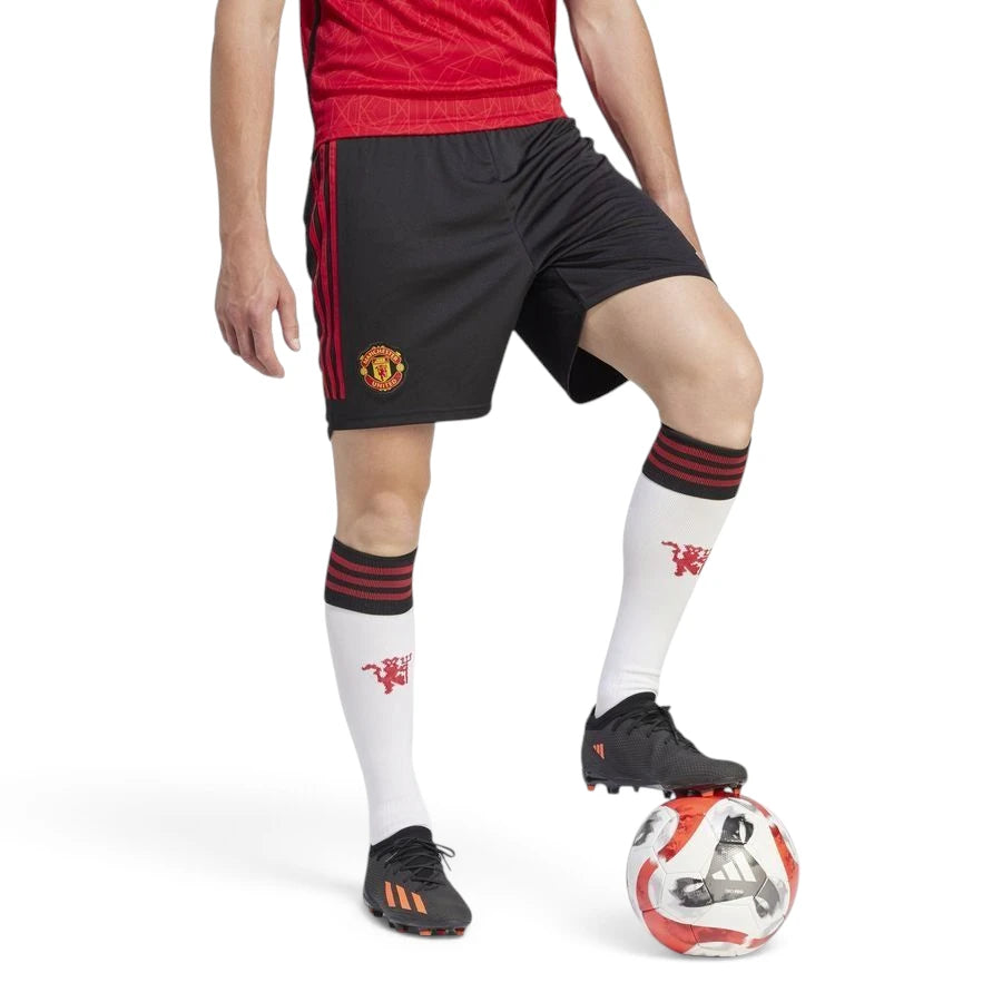 Studio action shot of the Manchester United 23/24 home shorts in black, highlighting the red 3-Stripes on the sides, the woven club crest, and the complete on-pitch kit look with white socks and football boots.