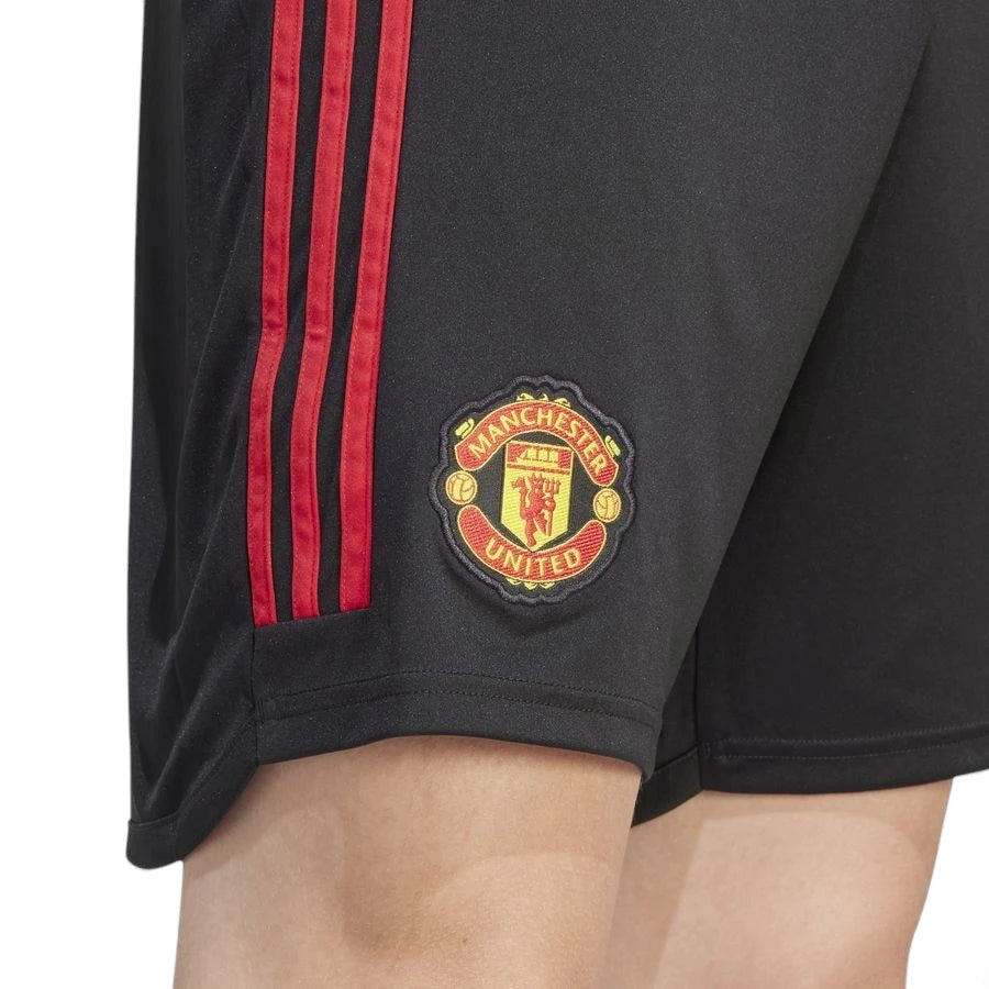 Macro close-up angle capturing the textured fabric weave, vibrant red 3-Stripes tape, and the precision embroidery of the Manchester United woven club badge.