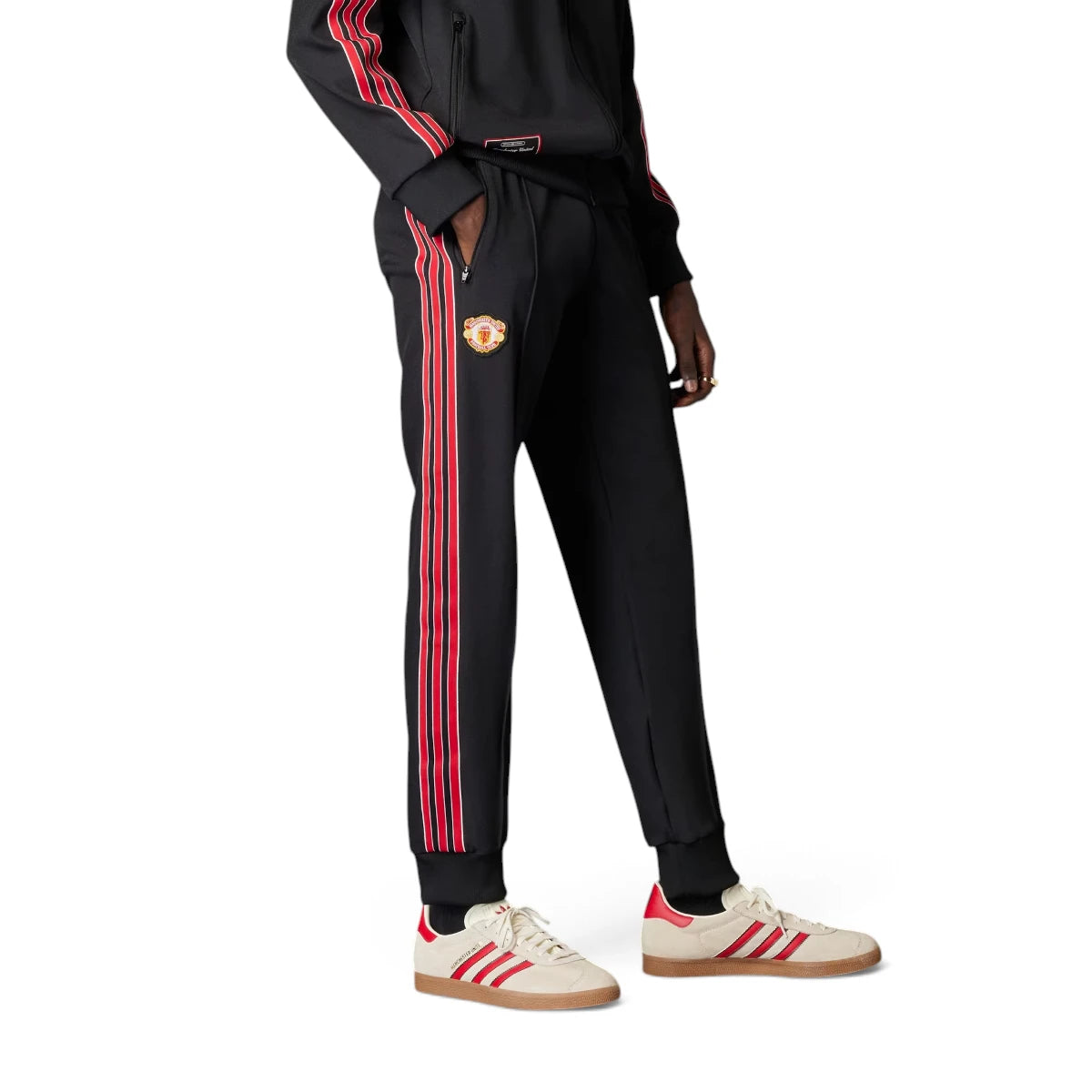 Studio side profile view of a model wearing the adidas Manchester United Terrace Icons Track Pants in black, highlighting the slim fit, red-and-white 3-Stripes taping, vintage Manchester United embroidered crest, front zip pockets, and ribbed cuffs. Main image