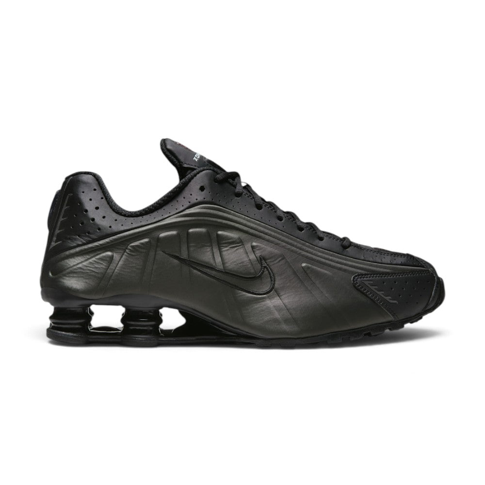 Lateral view showing the aerodynamic black synthetic casing, precision laser perforations along the midfoot, tonal Nike Swoosh badge, and the iconic four Bright Crimson Nike Shox columns compressed beneath the black TPU heel moderator plate. Main image