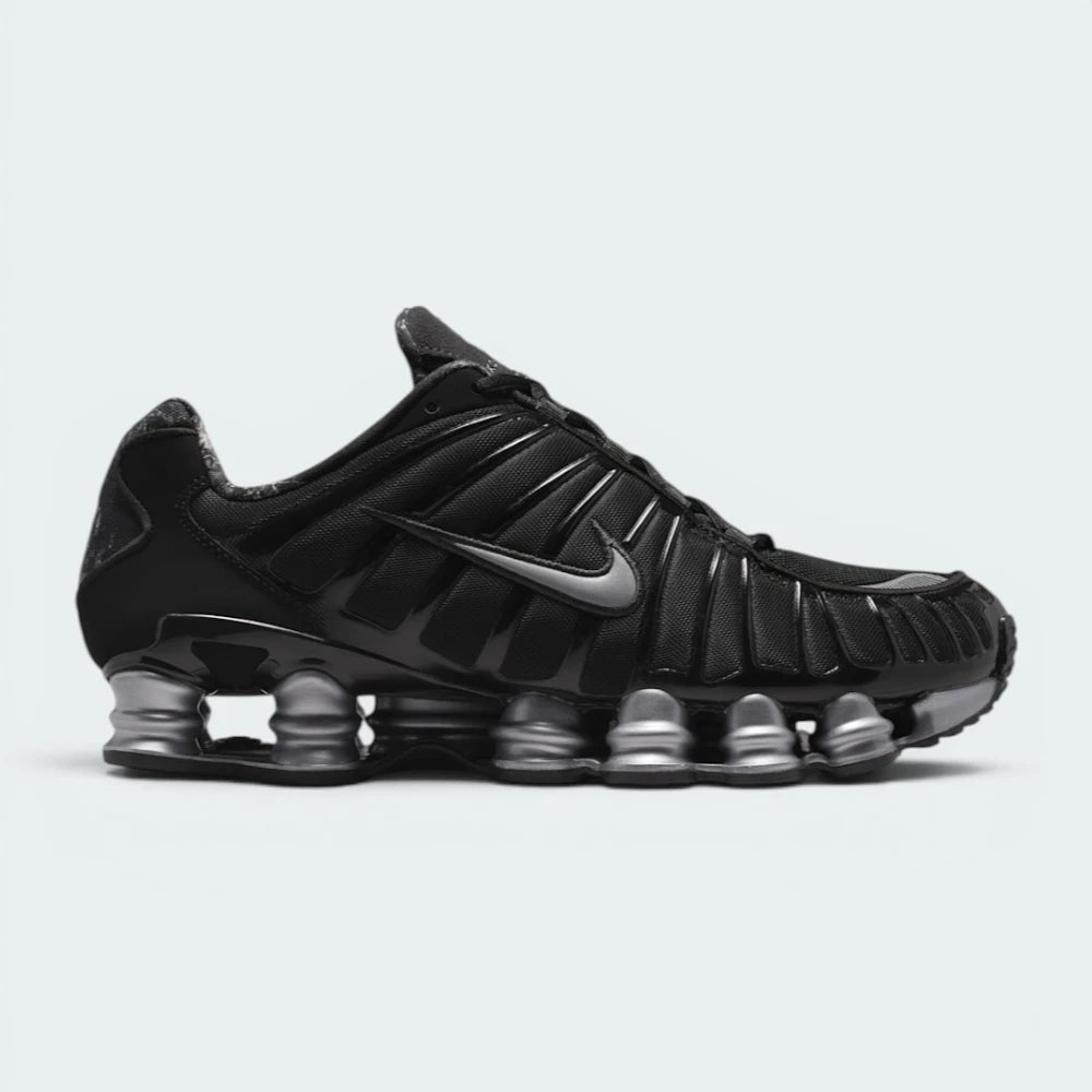Highlights the lateral profile showing the metallic silver Nike Swoosh, glossy black TPU cage, breathable black mesh quarter, metallic silver Shox columns extending the full length of the sole, and the subtle RealTree camo collar lining.