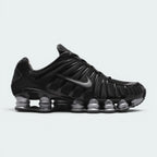 Highlights the lateral profile showing the metallic silver Nike Swoosh, glossy black TPU cage, breathable black mesh quarter, metallic silver Shox columns extending the full length of the sole, and the subtle RealTree camo collar lining.