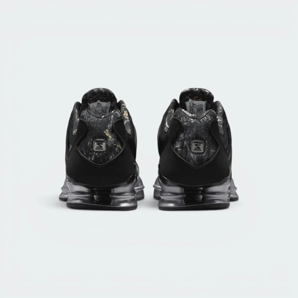 Focuses on the rear profile, highlighting the RealTree camo heel tab stamped with the metallic Shox "S" badge, sitting above the twin Shox heel columns and TPU stability plate.