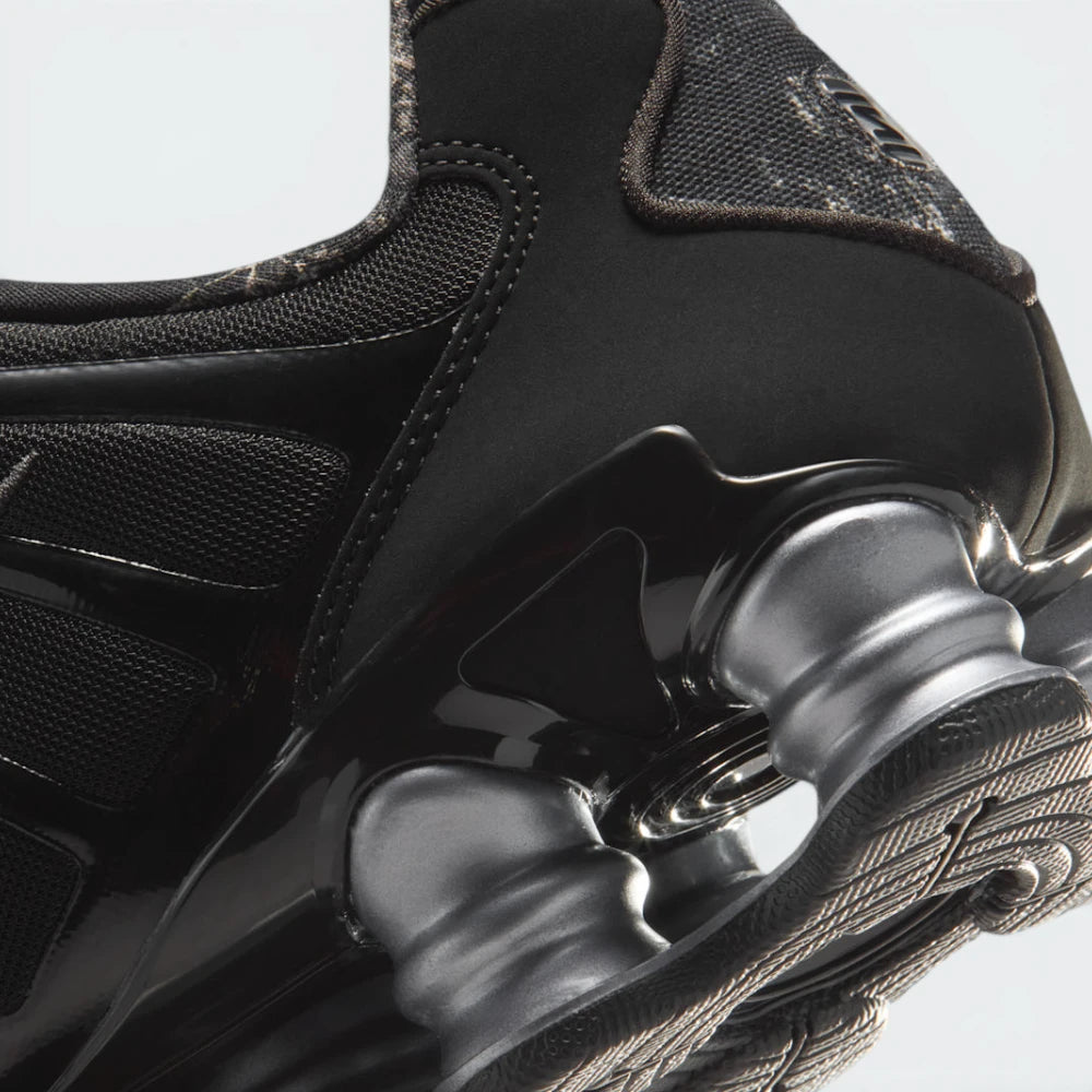 Highlights the "unreal" clean finish where the glossy black heel TPU moderator plate meets the metallic silver Shox columns and grooved black rubber outsole pods below.