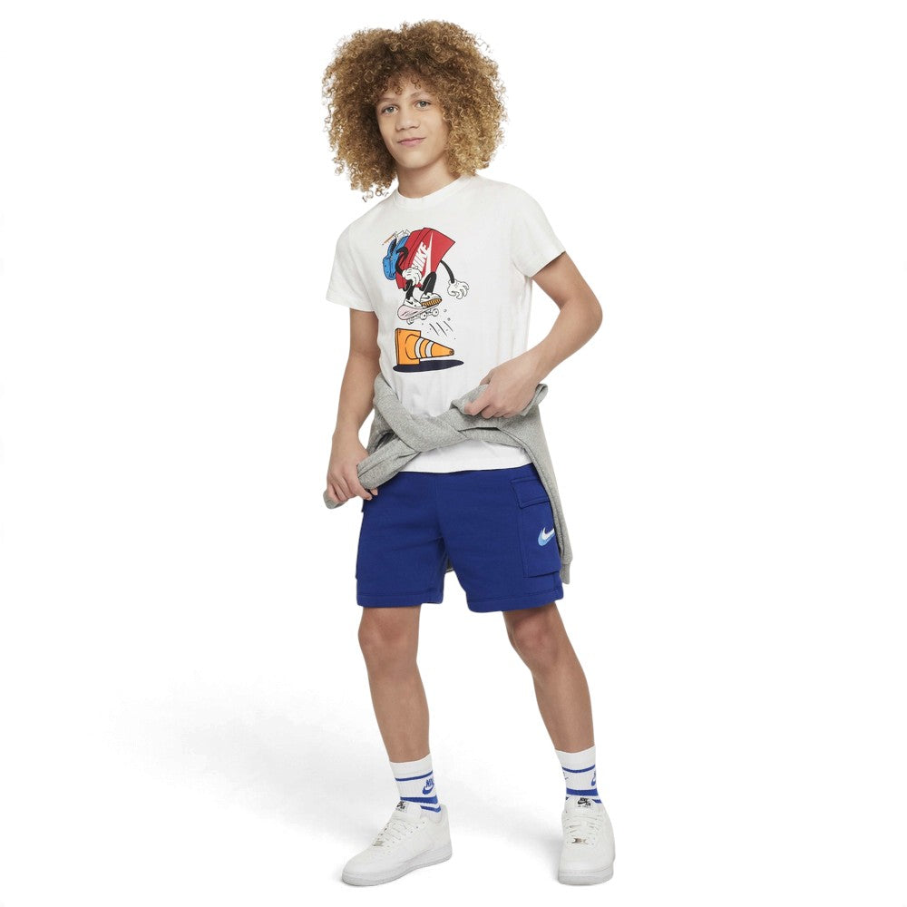 A child model wearing a white graphic t-shirt, blue shorts, a gray sweatshirt tied around the waist, white socks, and white sneakers, standing against a plain white studio background.