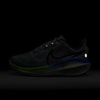 A dark Nike running shoe shown in low-light conditions against a black background, with visible green and blue accents on the midsole and a reflective white dot near the heel.