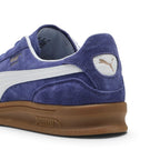 Close-up rear angle focusing on the Blue Crystal suede heel counter, white textured leather heel moustache tab stamped with a gilded metallic PUMA Cat logo, and the sculpted gum rubber heel bumper.